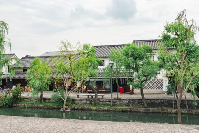 Traditional Japanese buildings with tiled roofs stand behind a canal, bordered by green trees and a stone pathway under a cloudy sky. Benches and a street lamp near the water complete this serene Kurashiki Travel guide scene.