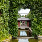 A modern building at Kurashiki Ivy Square, covered with dense green ivy, features a large rectangular opening framing a glass-walled walkway over a reflective water channel.