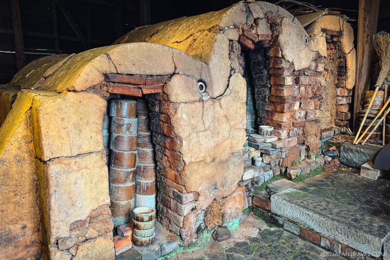 Two old brick and clay kilns with arched doorways, stacked with ceramic pots and containers inside, stand side by side on a stone floor in the dimly lit, rustic Kutaniyaki Kiln Museum.