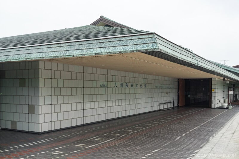 A modern building with a green-tiled sloped roof and light gray tiled walls, the Kyushu Ceramic Museum features Japanese text on the facade and a glass entrance door on the right. The walkway is paved with patterned tiles.