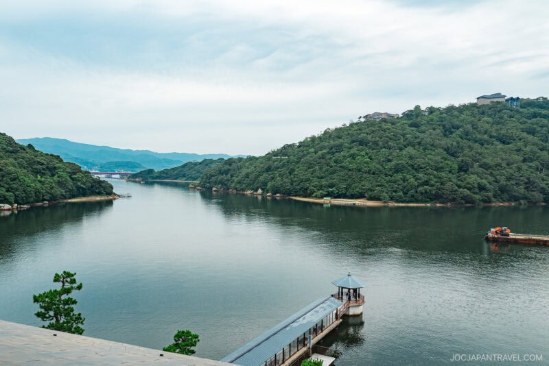A scenic view of tranquil Lake Hamana, surrounded by lush green hills, with a small pavilion on the shore and a boat on the water beneath a cloudy sky.
