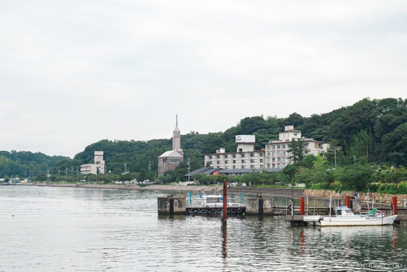 A calm Lake Hamana waterfront scene with several docked boats, a pier, and buildings along the shore. Green trees line the background under a cloudy sky.