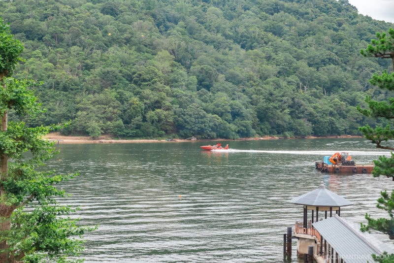 A red speedboat and a small work barge float on the calm waters of Lake Hamana, surrounded by lush green forested hills, with a wooden dock and pavilion nestled in the lower right corner.