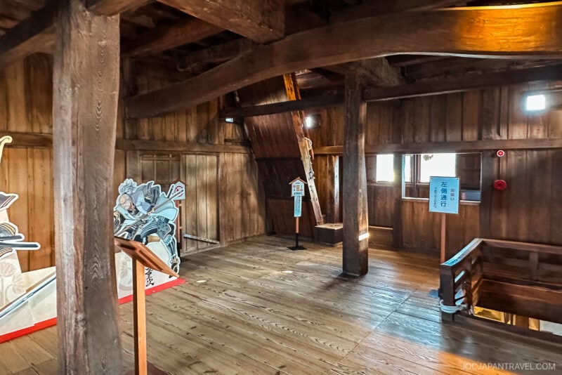 Interior of Maruoka Castle featuring exposed wooden beams, polished floors, natural light from small windows, and cutout samurai figures on display; informative signs enhance the historic ambiance of the room.