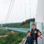 Mishima Skywalk (三島スカイウォーク) – A Thrilling Bridge with Mt. Fuji Views A man and two children stand smiling on Mishima Skywalk, a large suspension bridge with blue railings, surrounded by lush green hills and trees under a cloudy sky.