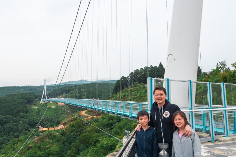 A man and two children stand smiling on Mishima Skywalk, a large suspension bridge with blue railings, surrounded by lush green hills and trees under a cloudy sky.