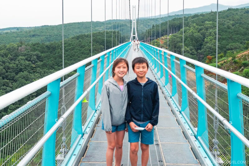 Two children, a girl and a boy, stand smiling on the blue Mishima Skywalk suspension bridge, surrounded by lush green trees and hills. The weather appears overcast, and the bridge stretches into the distance behind them.