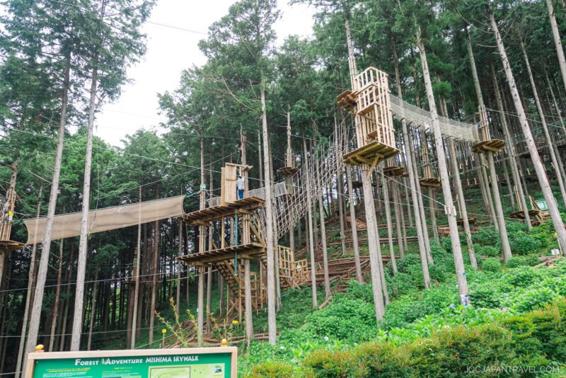 A tall wooden adventure course with rope bridges and platforms stands among lush green trees in a forest near Mishima Skywalk. A person in safety gear waits on a platform, while a green sign is visible in the foreground.