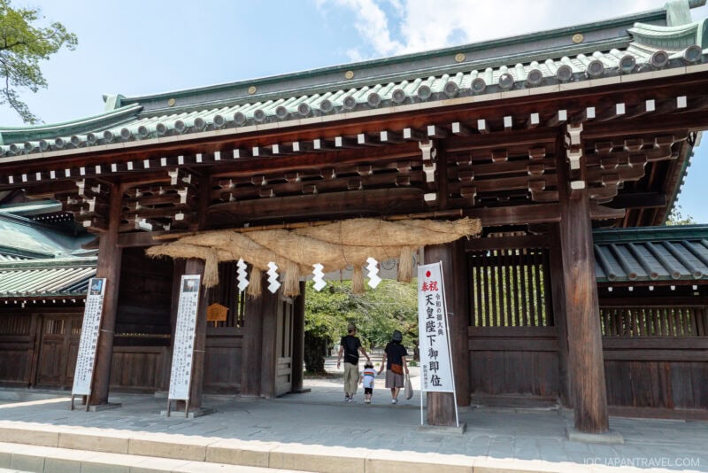 A traditional wooden Japanese shrine gate at Mishima Taisha Shrine features a large straw rope above the entrance; a family walks through, and informational signs are posted on either side of the gate.