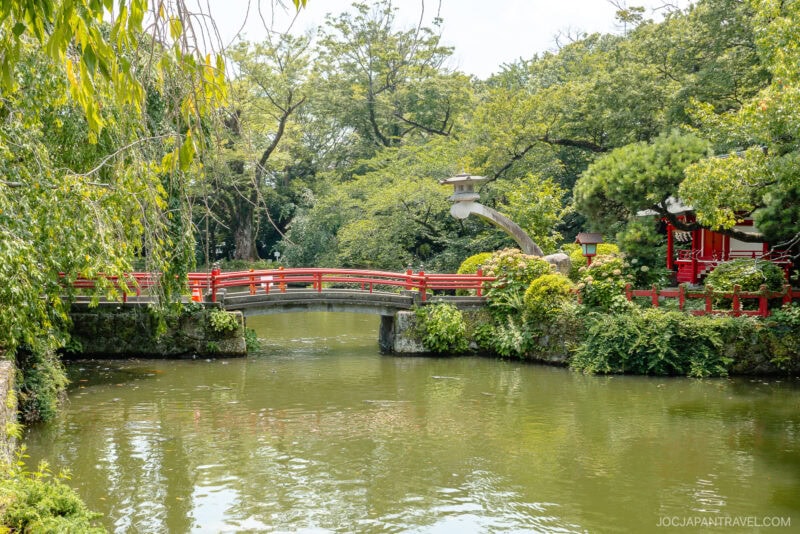 A red arched bridge crosses over a peaceful pond surrounded by lush greenery and trees in a traditional Japanese garden near Mishima Taisha Shrine. A stone lantern and a red building are partially visible on the right.