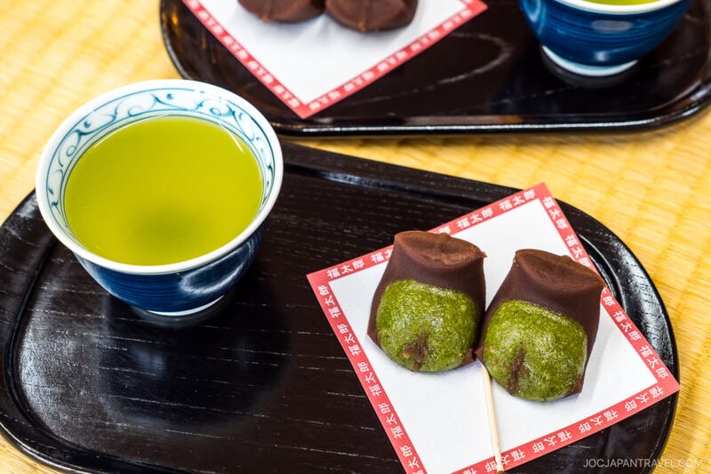 A cup of green tea and two pieces of Japanese wagashi sweets with green mochi and red bean paste are served on a black tray, evoking the serene atmosphere near Mishima Taisha Shrine. Another similar tray is seen in the background.