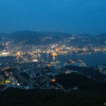 Mt. Inasayama Observatory (稲佐山山頂展望台) – Nagasaki’s Best Panoramic Lookout Aerial view of Nagasaki, Japan at dusk, with city lights illuminating the buildings, streets, and harbor, surrounded by hills and Mt. Inasayama beneath a deep blue sky.