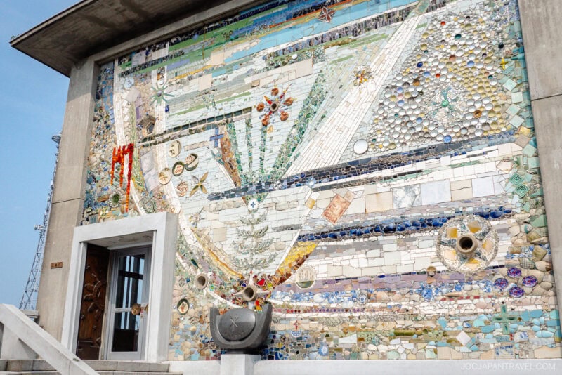 A building facade decorated with a colorful mosaic of tiles, glass, and ceramics, featuring abstract designs and various objects, reminiscent of the vibrant Museum of the 26 Martyrs. The entrance has a small set of steps and a modern-style door.