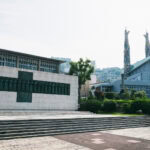 A modern memorial with bronze relief sculptures stands near the Museum of the 26 Martyrs, alongside a building with tall, decorated columns, surrounded by green landscaping and city buildings beneath a bright sky.