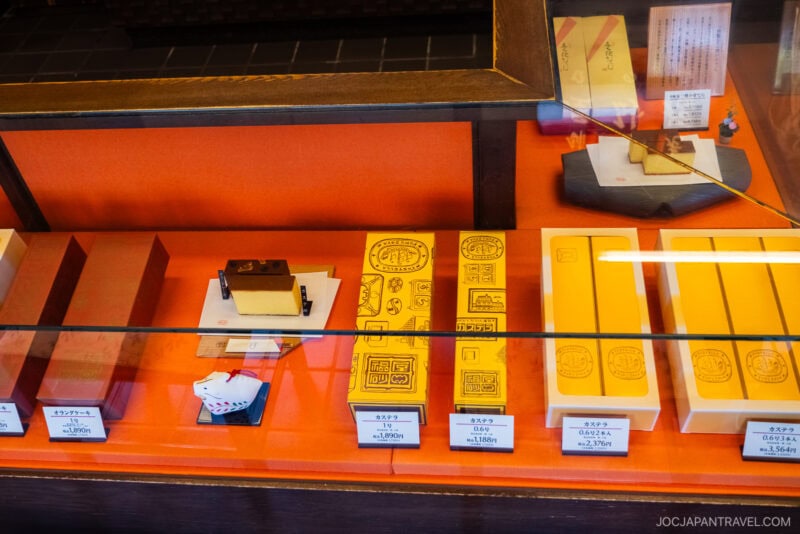 A display case shows neatly arranged Nagasaki Castella Cakes or sweets in yellow packaging with illustrated designs, along with a small ceramic cow figurine and price tags in Japanese yen.