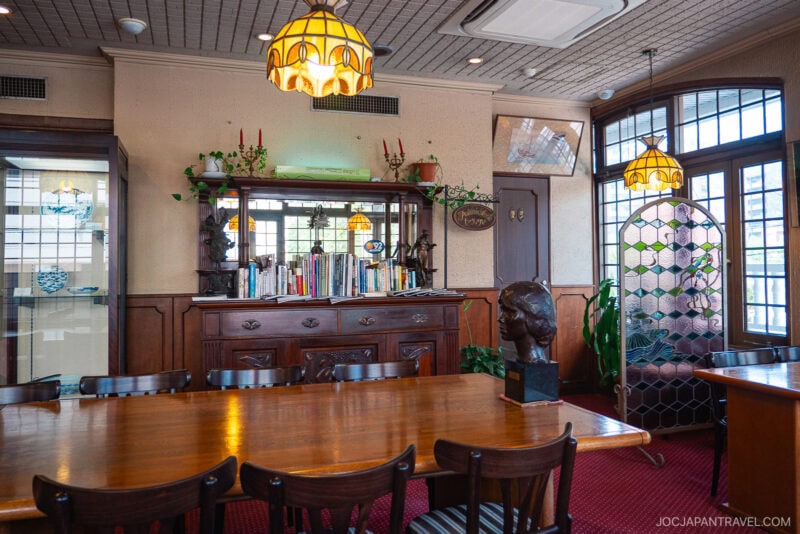 A cozy, vintage-style dining room with a wooden table and chairs, bookshelves, decorative lamps, a plant, and a bust sculpture. Large windows let in natural light; Nagasaki Castella Cake is beautifully displayed beneath stained glass.