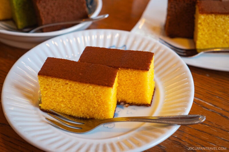 Two rectangular slices of golden yellow Nagasaki Castella Cake with brown tops are served on a white plate, each with a small fork beside them. Other plates featuring this classic cake are visible in the background.