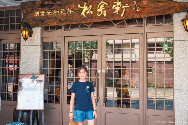 A young woman in a navy shirt and shorts stands smiling in front of a traditional Japanese restaurant with wooden doors and a large wooden sign above the entrance, where Nagasaki Castella Cake is a specialty.
