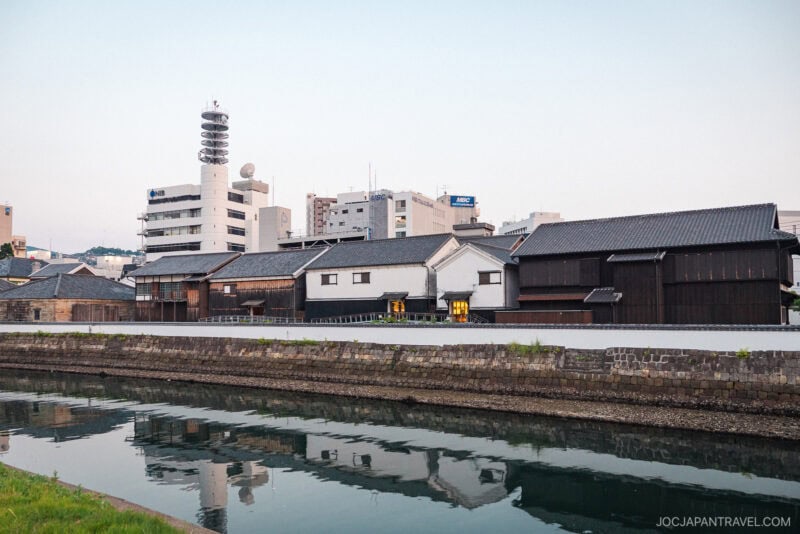Row of traditional Japanese warehouses with white walls and dark roofs beside a calm canal in Dejima, Nagasaki, with modern office buildings and a communications tower in the background.