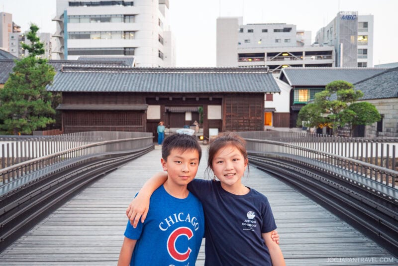 Two children stand on a wooden bridge in Dejima, Nagasaki, smiling with their arms around each other. Modern buildings and traditional Japanese architecture create a striking backdrop.