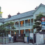 Dejima Nagasaki (出島) – Walking Through Japan’s Age of Isolation A two-story Western-style building in Dejima, Nagasaki, with a clock tower, white wooden walls, blue trim, balconies, and multiple signs in front—surrounded by trees and a fence at dusk.
