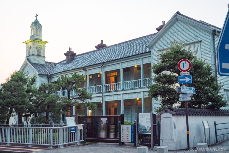 A two-story Western-style building in Dejima, Nagasaki, with a clock tower, white wooden walls, blue trim, balconies, and multiple signs in front—surrounded by trees and a fence at dusk.