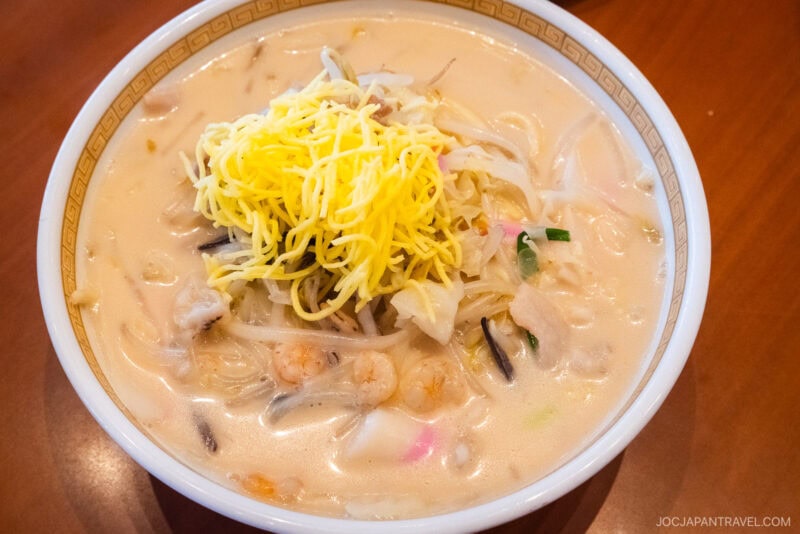 A bowl of creamy white soup filled with Nagasaki noodles, shrimp, vegetables, and topped with a small pile of shredded yellow egg, served on a brown table.