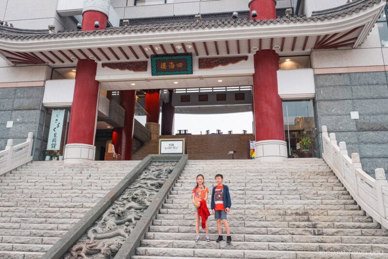 Two children stand on wide stone steps in front of a large building with red columns and ornate decorations, featuring dragon carvings on the stair rail. The scene evokes the traditional East Asian style found in places famous for nagasaki noodles.