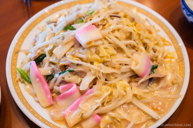 A plate of champon, a Japanese noodle dish known for its nagasaki noodles, topped with vegetables, pink-and-white kamaboko fish cakes, and pieces of meat in a creamy, savory broth.