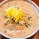 Nagasaki Noodles – Champon and Sara Udon A bowl of creamy white soup filled with Nagasaki noodles, shrimp, vegetables, and topped with a small pile of shredded yellow egg, served on a brown table.