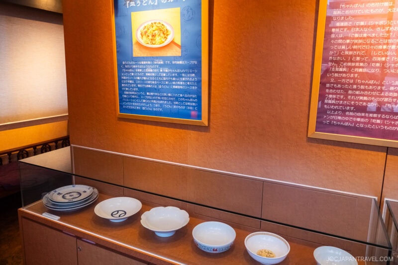 A display of empty bowls and plates on a countertop beneath two large information panels with Japanese text and images, highlighting the story of Nagasaki noodles in a well-lit indoor setting.