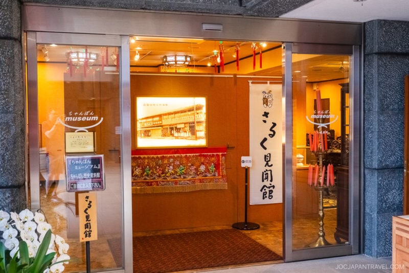 Entrance to a museum with glass doors, Japanese banners, floral decorations, and warm lighting. A sign with “Amuseum” is visible, and the interior displays traditional Japanese ornaments, framed art, and nods to Nagasaki noodles culture.