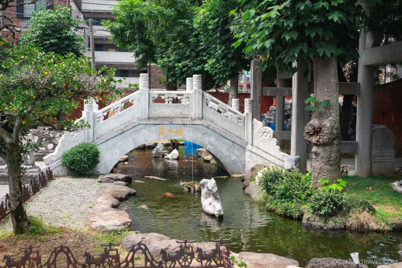 A traditional white stone bridge crosses a small pond with rocks and koi fish, surrounded by lush trees and greenery at Nagasaki Koshibyo Confucius Shrine. Urban buildings rise in the background, blending historic charm with city life.