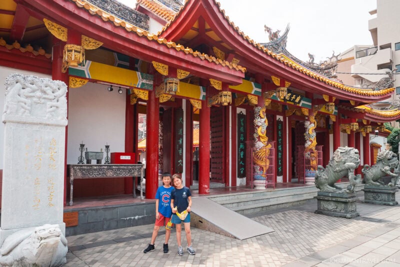 Two children stand smiling in front of Nagasaki Koshibyo Confucius Shrine, a traditional Chinese temple with ornate red columns, colorful decorations, dragon statues, lanterns, and intricate architectural details.