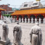 Nagasaki Travel Guide (長崎市) – History, Culture, and Nature in Kyushu Rows of stone statues in traditional robes stand in the courtyard of Nagasaki Koshibyo Confucius Shrine, with bright orange-tiled roofs and urban buildings visible in the background.