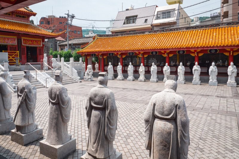 Rows of stone statues in traditional robes stand in the courtyard of Nagasaki Koshibyo Confucius Shrine, with bright orange-tiled roofs and urban buildings visible in the background.
