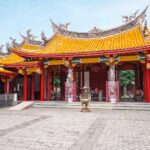 Nagasaki Koshibyo Confucius Shrine (長崎孔子廟) The Nagasaki Koshibyo Confucius Shrine is a traditional Chinese temple with ornate yellow-tiled roofs, red columns, and decorative dragon sculptures. The courtyard features stone statues and a large incense burner at the center.