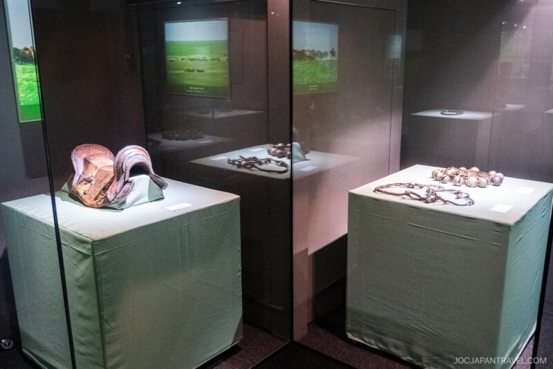 Museum display with two glass cases showing antique horse riding gear and metal objects, including a saddle on the left and tack on the right, inspired by artifacts at Nagasaki Koshibyo Confucius Shrine, with photos of grassy fields in the background.