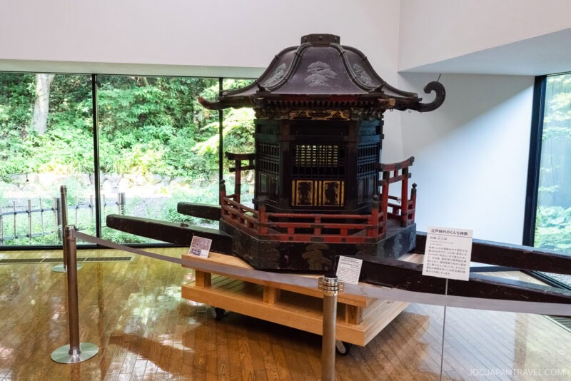 A traditional Japanese palanquin with a curved roof and wooden railings is displayed indoors on a platform at the Nagasaki Museum of History and Culture, with signs providing information and a large window revealing greenery outside.