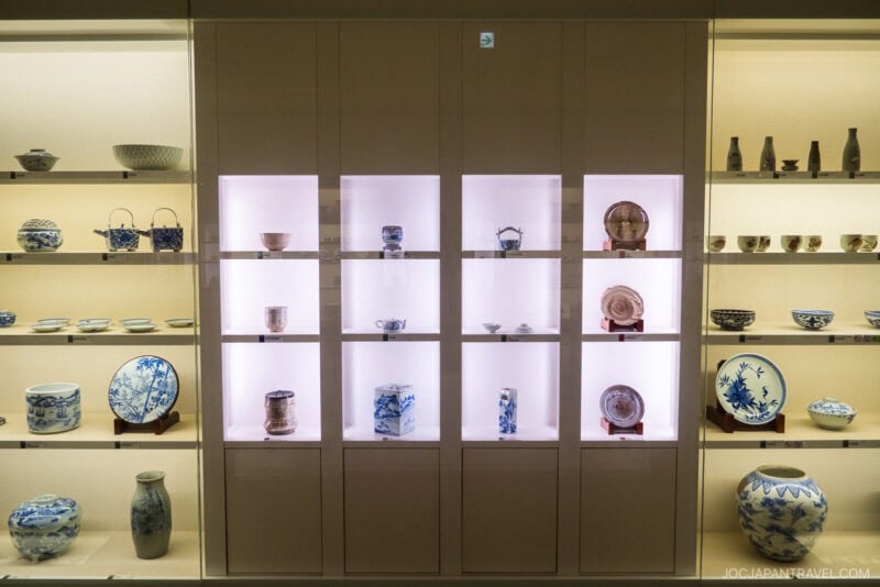 A display case with illuminated shelves showcases various traditional Japanese ceramic bowls, plates, and vases in blue and white patterns, all arranged neatly in the Nagasaki Museum of History and Culture.