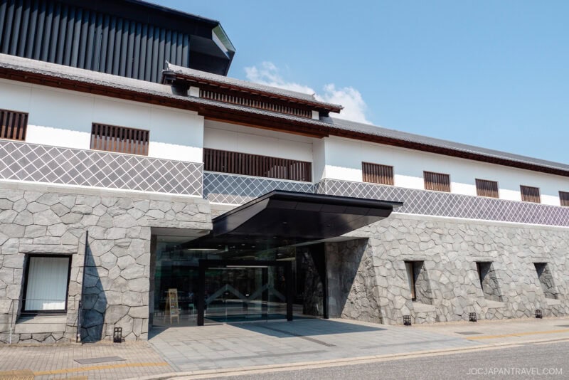 Modern building with traditional Japanese architectural elements, including white walls, lattice patterns, and a stone base. The Nagasaki Museum of History and Culture features a glass entrance with a canopy under a clear blue sky.