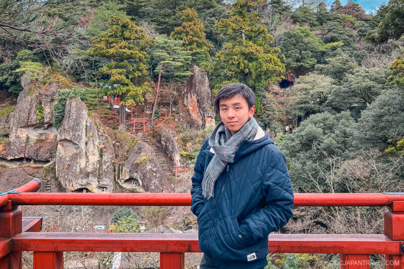 A young person in a blue jacket and scarf stands on a red railing with a scenic, forested mountain and rocky cliffs in the background, featuring Natadera Temple’s Torii gate and shrines nestled among the green trees.