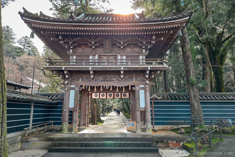 A traditional wooden Japanese temple gate, like those at Natadera Temple, stands at the entrance to a peaceful pathway lined with tall trees, sunlight streaming through the structure and a few boxes placed near the gate.