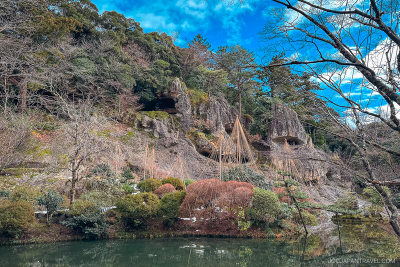 A peaceful Japanese garden at Natadera Temple features a pond in the foreground, trees, bushes, and rocky cliffs. Some plants are protected with straw coverings, and the sky is partly cloudy with patches of blue.