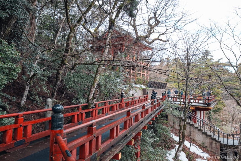 A red wooden bridge with railings winds through a forested hillside, leading to the historic Natadera Temple surrounded by trees and patches of snow. Several people are walking along the bridge and stairs toward the temple.