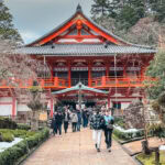 Natadera Temple (那谷寺) – Sacred Caves and Scenic Beauty in Kaga Visitors walk along a stone path toward Natadera Temple, its red and white facade nestled among trees and surrounded by patches of snow.