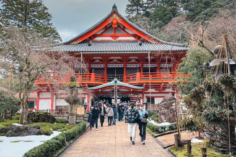 Visitors walk along a stone path toward Natadera Temple, its red and white facade nestled among trees and surrounded by patches of snow.