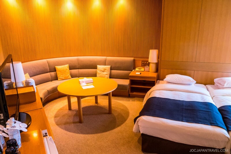 A cozy New Furano Prince Hotel room with two single beds, a round wooden table, a curved sectional sofa with cushions, and warm lighting on wood-paneled walls. A TV and amenities are on a small desk nearby.
