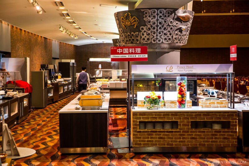At the New Furano Prince Hotel, a buffet area features a Chinese cuisine station with food displays under glass, decorative brickwork, and a large ornament overhead. A person in a purple shirt walks by, and the sign reads Chinese cuisine in English and Chinese.