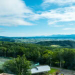 New Furano Prince Hotel (新富良野プリンスホテル) – A Comfortable Stay Near the Flower Fields View of a lush green landscape with tennis courts and buildings in the foreground, dense forests, rolling hills, and an expansive blue sky with scattered clouds—likely taken from the New Furano Prince Hotel’s elevated vantage point.